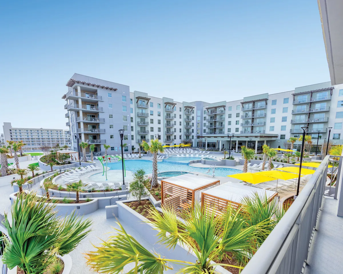 The photo captures a stylish pool area dotted with bright yellow umbrellas, modern resort architecture, and palm trees. The scene is lively, with guests swimming, relaxing, and enjoying social gatherings in the sun-drenched space.