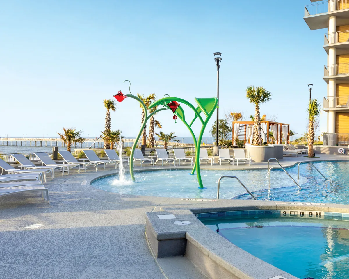 This image features a vibrant pool area, complete with a water play structure, lounge chairs, palm trees, and a sunny sky. Children are seen playing and splashing in the water, while adults relax by the poolside, creating a lively, family-friendly atmosphere.