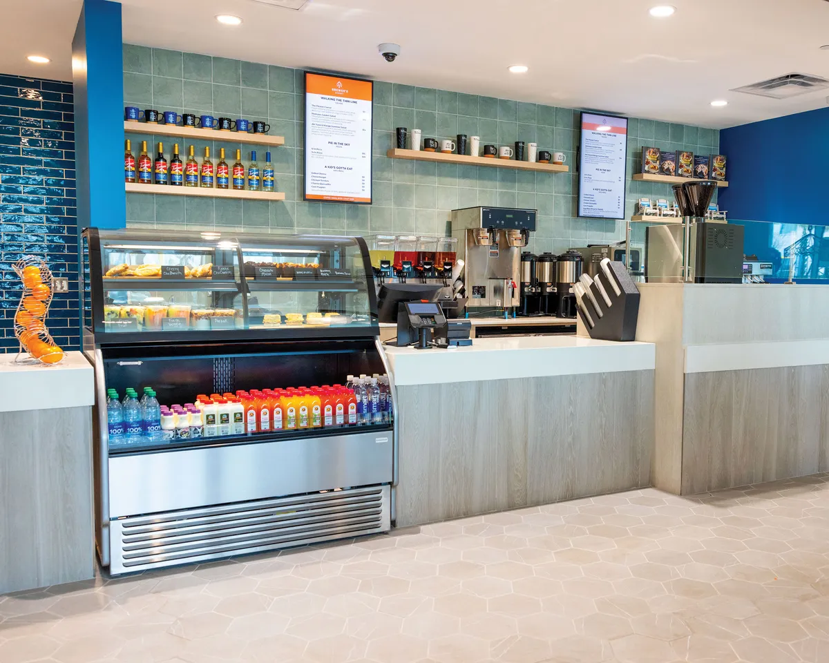 Inside a welcoming café, friends gather around a counter displaying tempting pastries and a beverage cooler, while others chat and laugh near the coffee station. The space is bright and casual, with menu boards overhead and a relaxed, social vibe throughout.