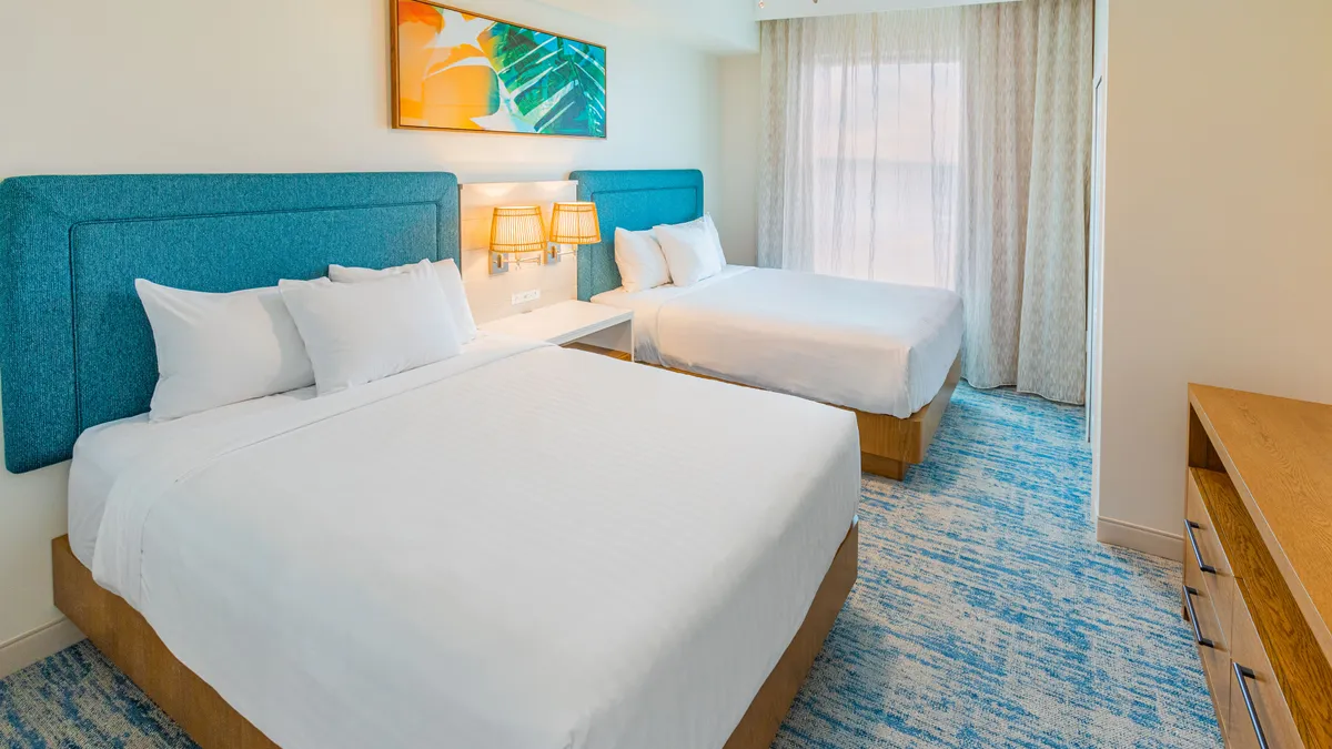 A comfortable and inviting second bedroom featuring two queen beds dressed in crisp white linens. Colorful artwork adds personality, while a large window with sheer curtains floods the space with natural light. The atmosphere is tranquil, offering an ideal environment for families or groups to rest and reconnect.