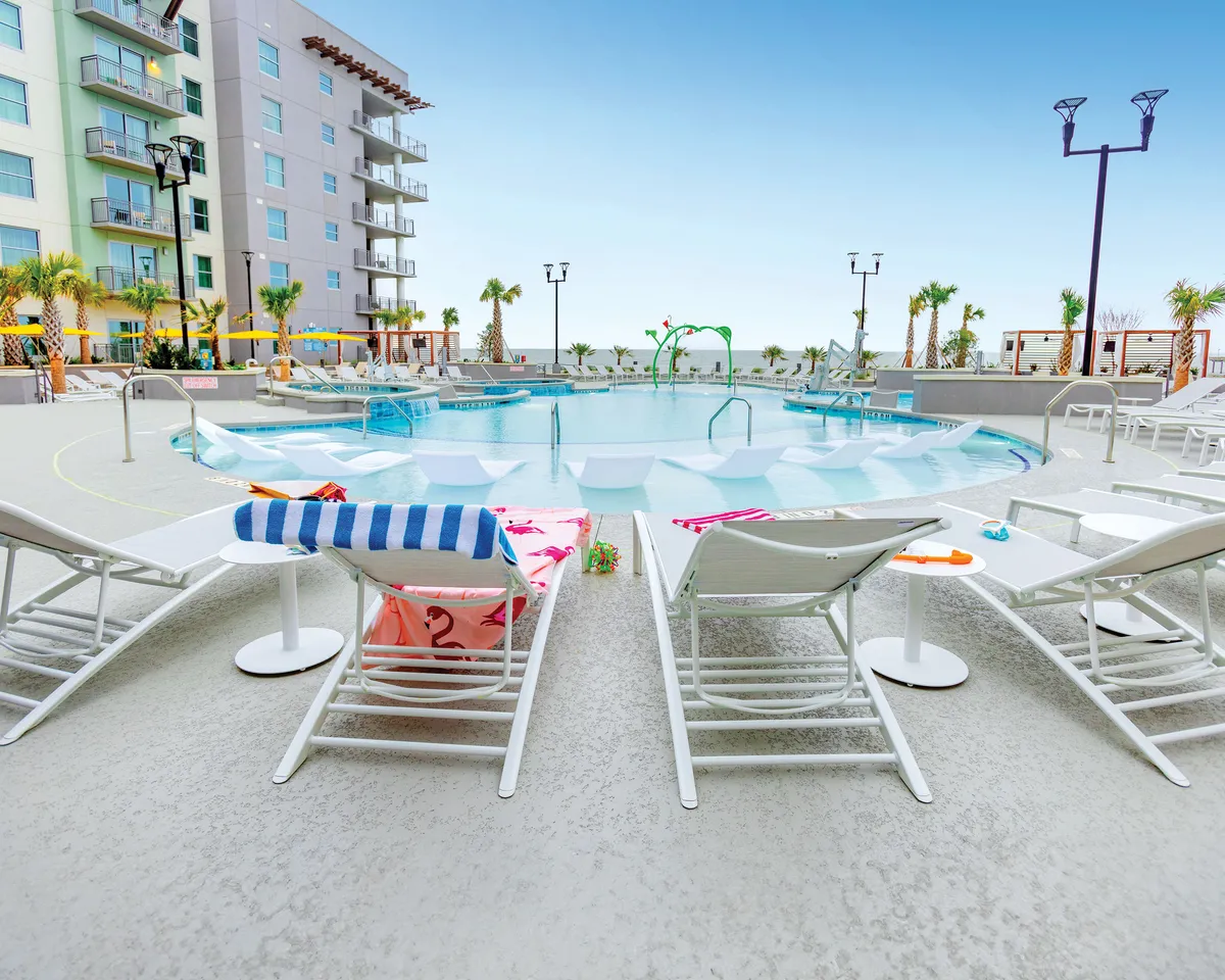 A spacious swimming pool is framed by lounge chairs with colorful towels, swaying palm trees, and the balconies of the resort building in the background. The setting is relaxed, with families and groups enjoying the sun and water together.