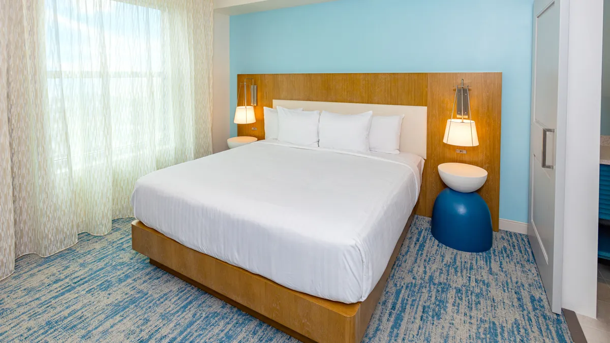 A serene master bedroom dominated by a plush king bed, flanked by nightstands and soft window curtains. The room features calming coastal hues and thick carpeting, inviting rest and relaxation after a day at the beach. Natural light filters in, creating a soothing ambiance ideal for recharging.