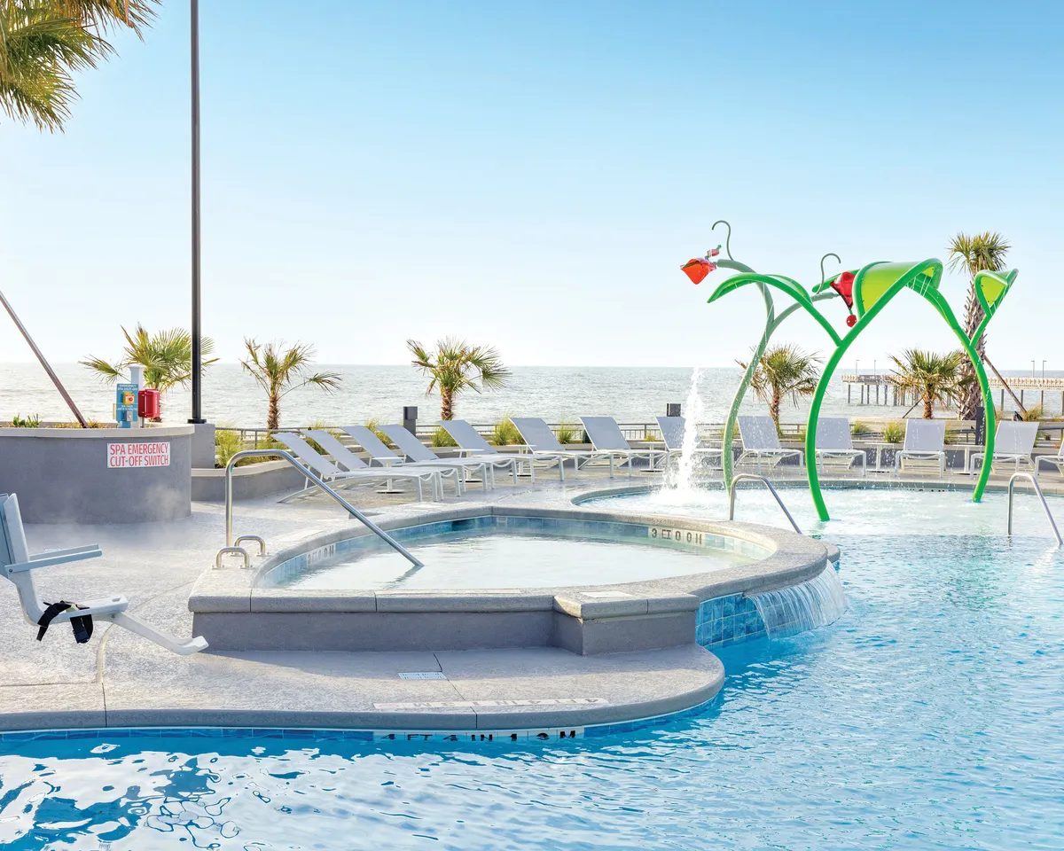 This vibrant exterior scene captures a large swimming pool with playful water features and colorful lounge chairs, all framed by a sweeping sea view. Children are seen enjoying the splash structures while families relax poolside, evoking energy and fun. The setting sun casts a golden glow, enhancing the inviting atmosphere and highlighting both family-friendly amenities and the resort's prime beachside location.