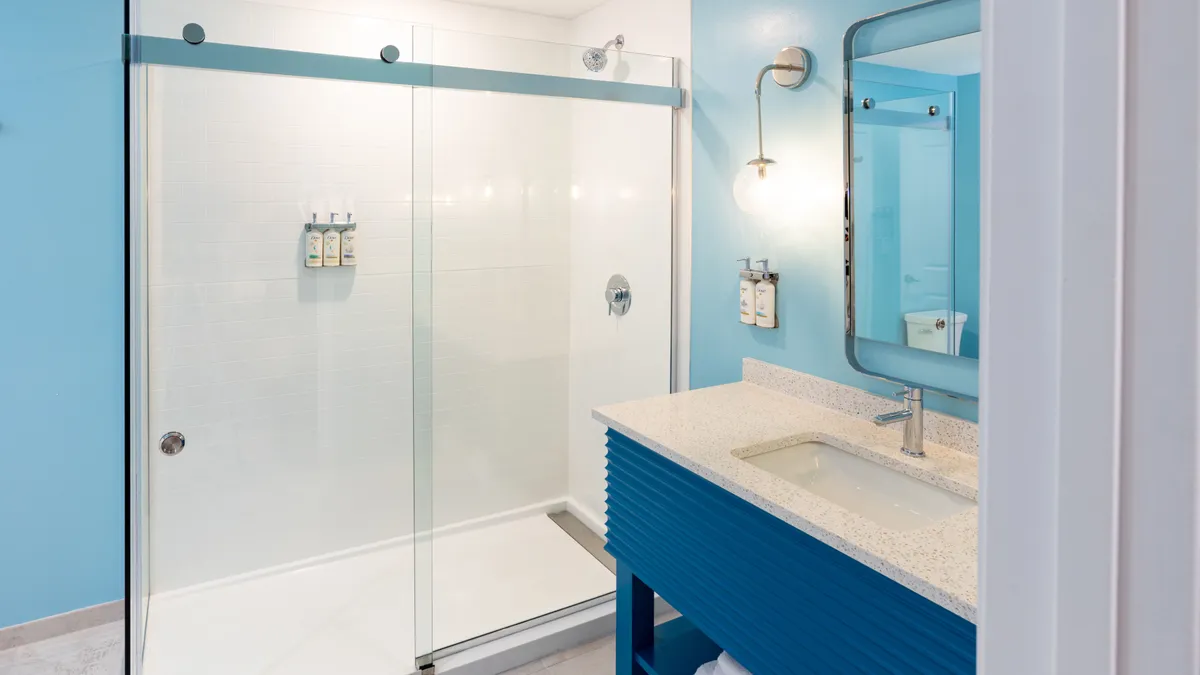A bright, modern bathroom with a glass walk-in shower, blue vanity, and ample lighting. The spa-like ambiance encourages relaxation and self-care, while practical design ensures accessibility and comfort for all guests.