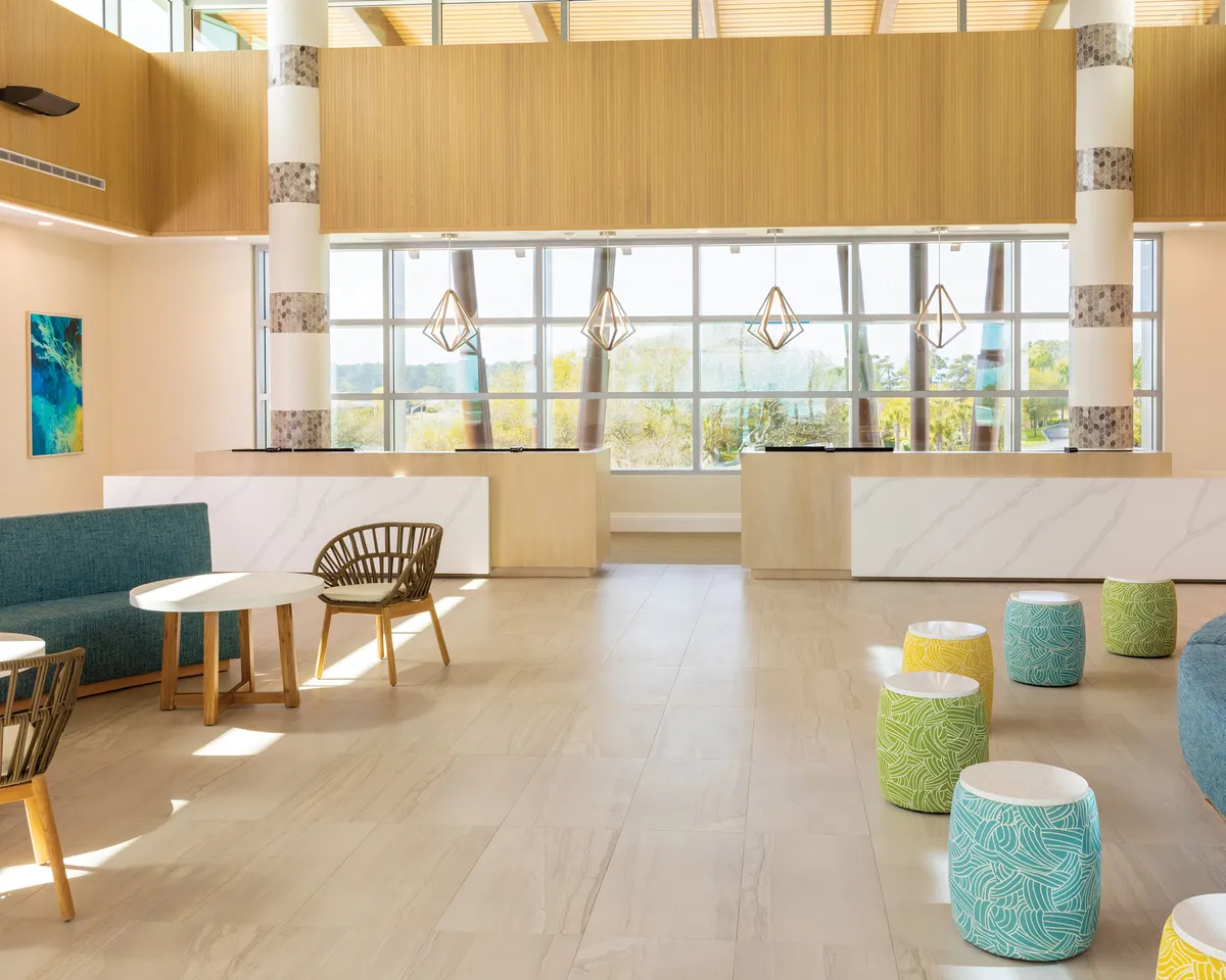 A bright interior space with large windows streaming in natural light, colorful stools around a modern reception area, and contemporary furniture. The lively, inviting mood encourages social gatherings and creates a vibrant backdrop for group activities or relaxed moments.