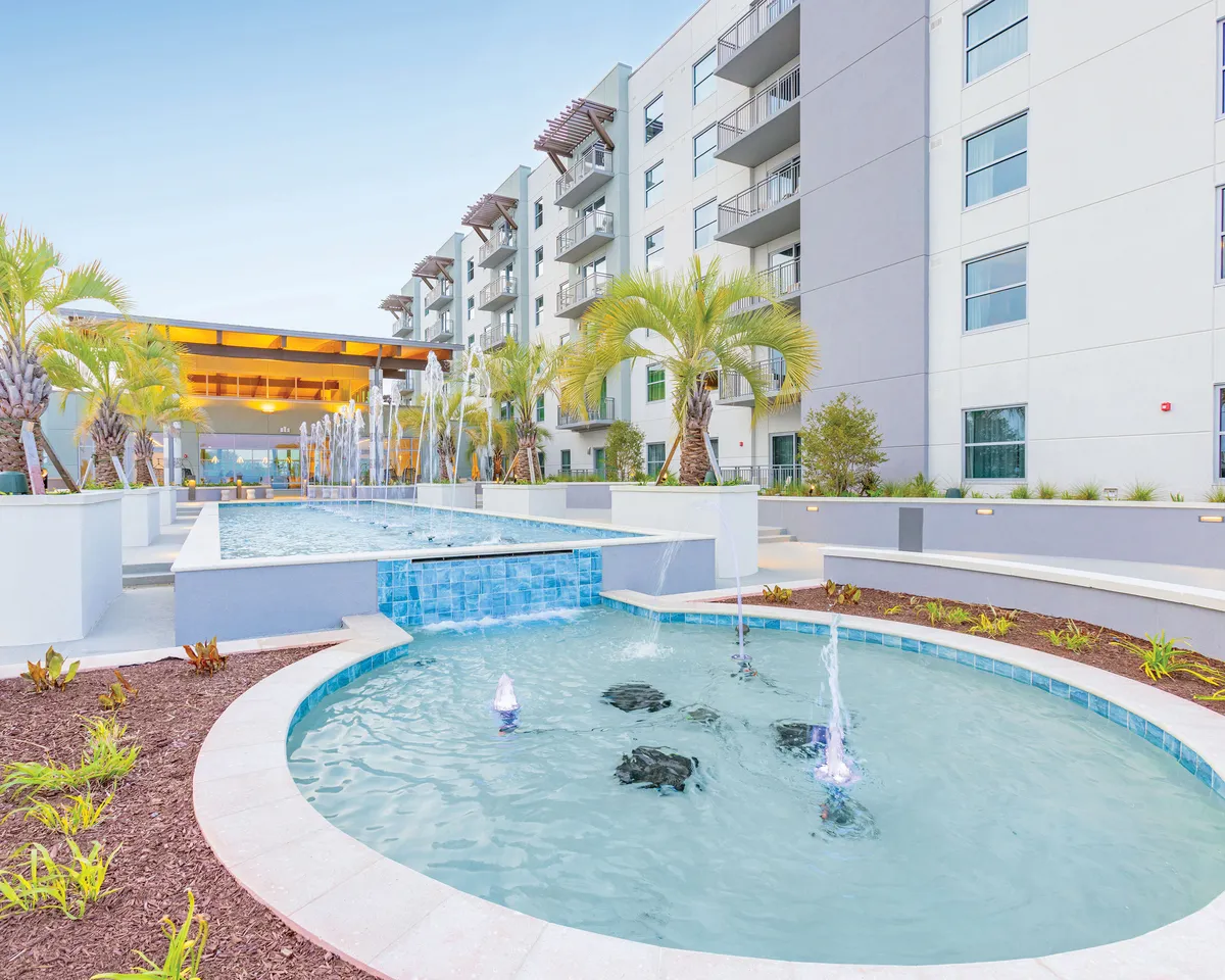 This image features the exterior pool area of the resort at sunset, with inviting pools accented by fountains, lush landscaped spaces, and a modern building facade. Groups are seen relaxing by the poolside, mingling and enjoying the serene ambiance, which hints at fun social gatherings and evening events.