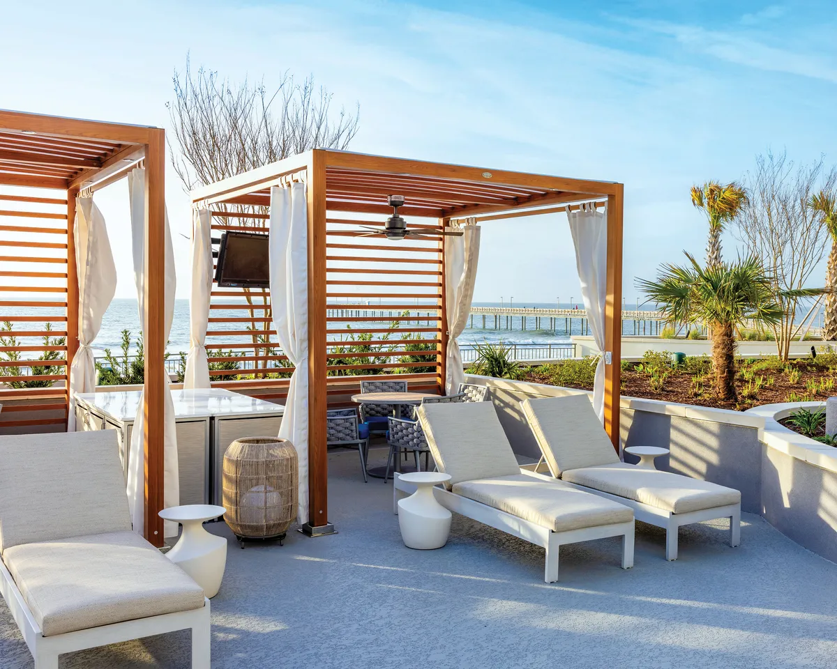 This image features a serene outdoor balcony or terrace area with wooden cabanas, sun loungers, a view of the water, lush greenery, and an outdoor dining area. The setting suggests tranquil afternoons and intimate gatherings with loved ones while enjoying the refreshing sea breeze.