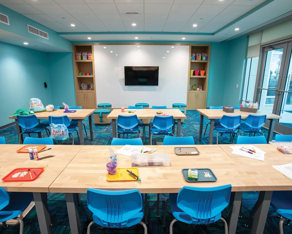 A lively activity room outfitted with tables covered in craft supplies, colorful chairs, shelves lined with creative items, and a large television. The space is designed for arts, crafts, and group workshops, offering a cheerful setting for shared experiences across generations.